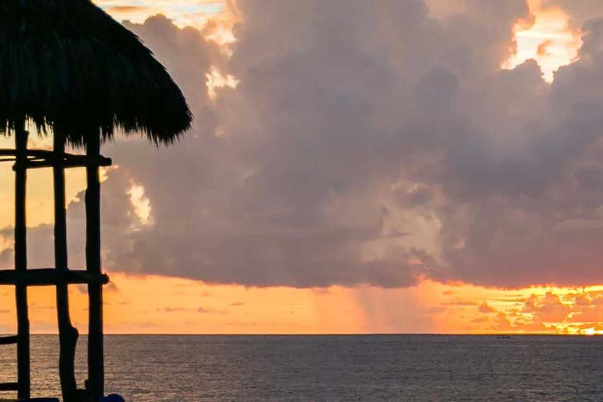 A thatched palapa in silhouette against an orange sunset and heavy clouds over the ocean.