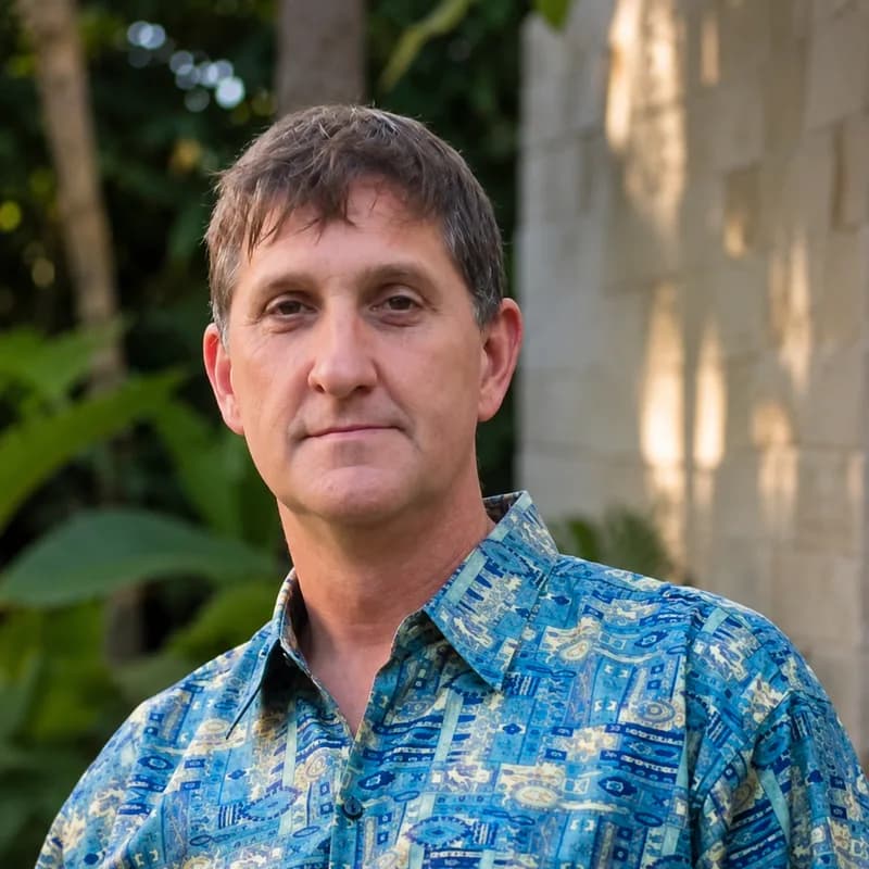 A man with short brown hair in a blue patterned shirt, with green leaves behind him.