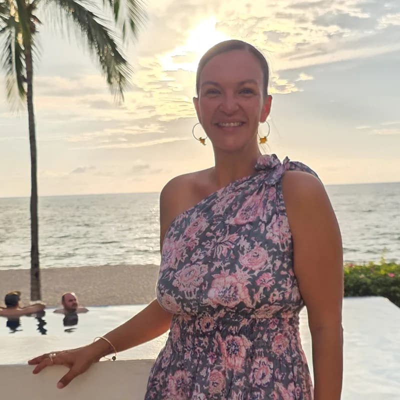 A smiling woman in a floral one-shoulder dress, with a sunset sky behind her.