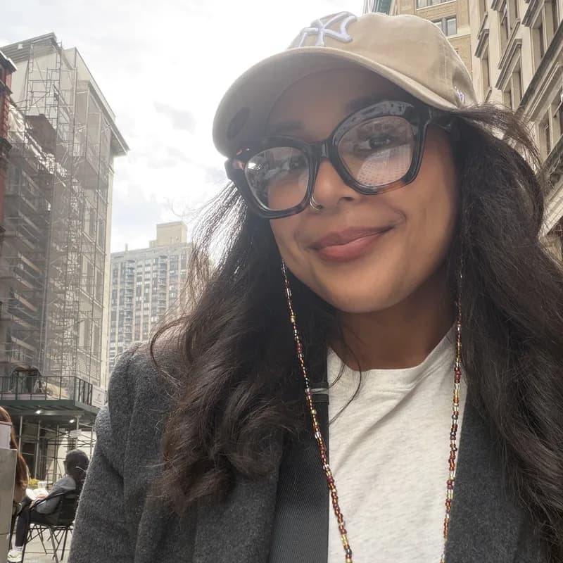 A smiling woman in a tan cap and dark-framed glasses on a city street.
